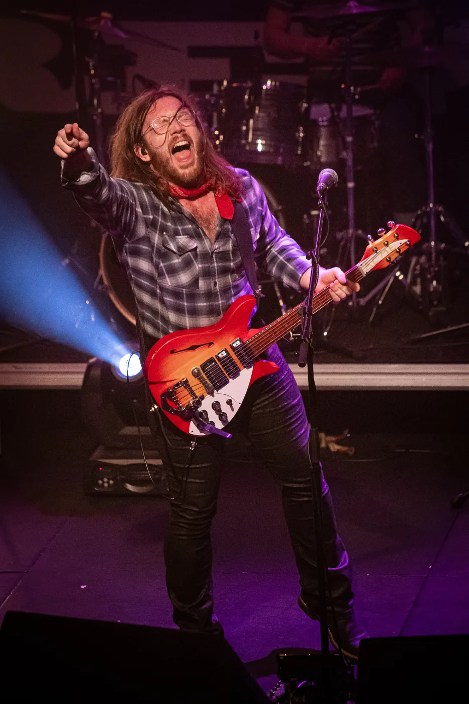 A guitarist in a checked shirt and red neckerchief points at the audience while singing into a microphone on stage