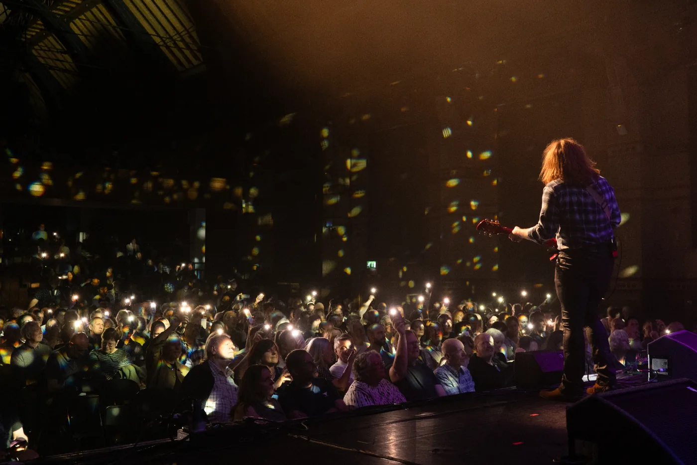 View from the side of a stage towards an audience holding up lit phones, with confetti falling and a guitarist silhouetted in the foreground