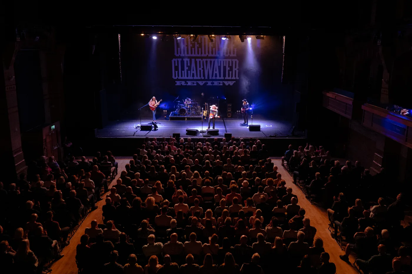 A full theatre auditorium seen from the balcony, with four musicians on a lit stage under a large Creedence Clearwater Review backdrop